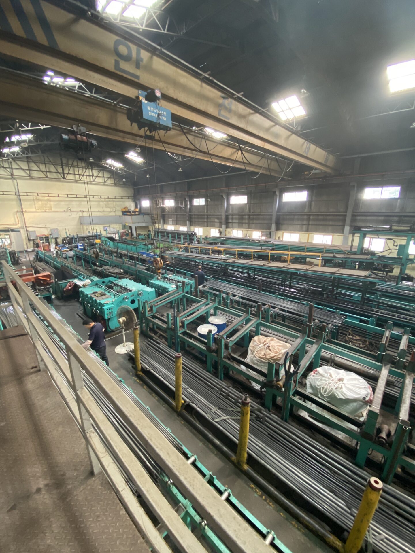 An industrial interior with a person working among large machinery, conveyor belts, and metal parts in a well-lit facility with an overhead crane.