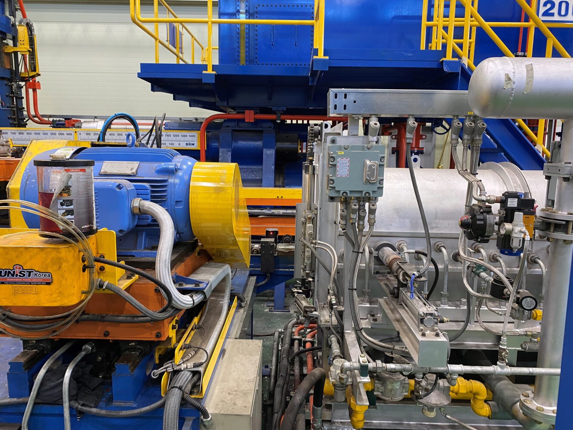 The image shows industrial equipment with pipelines, valves, and motors inside a facility with blue and yellow machinery and safety railings.