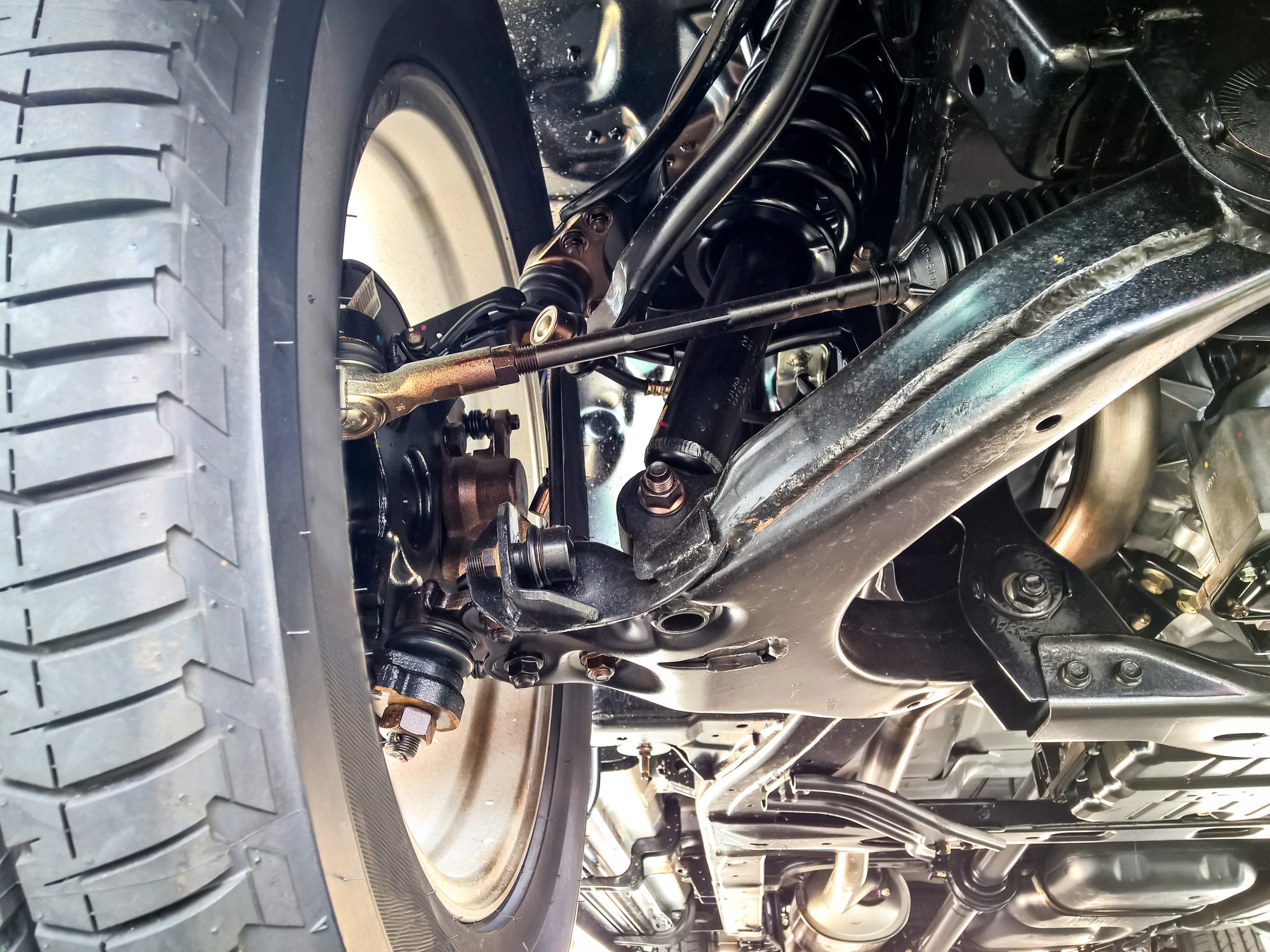 The image shows the undercarriage of a vehicle, focusing on the suspension system, wheel, and some mechanical components, with visible axles, joints, and springs.