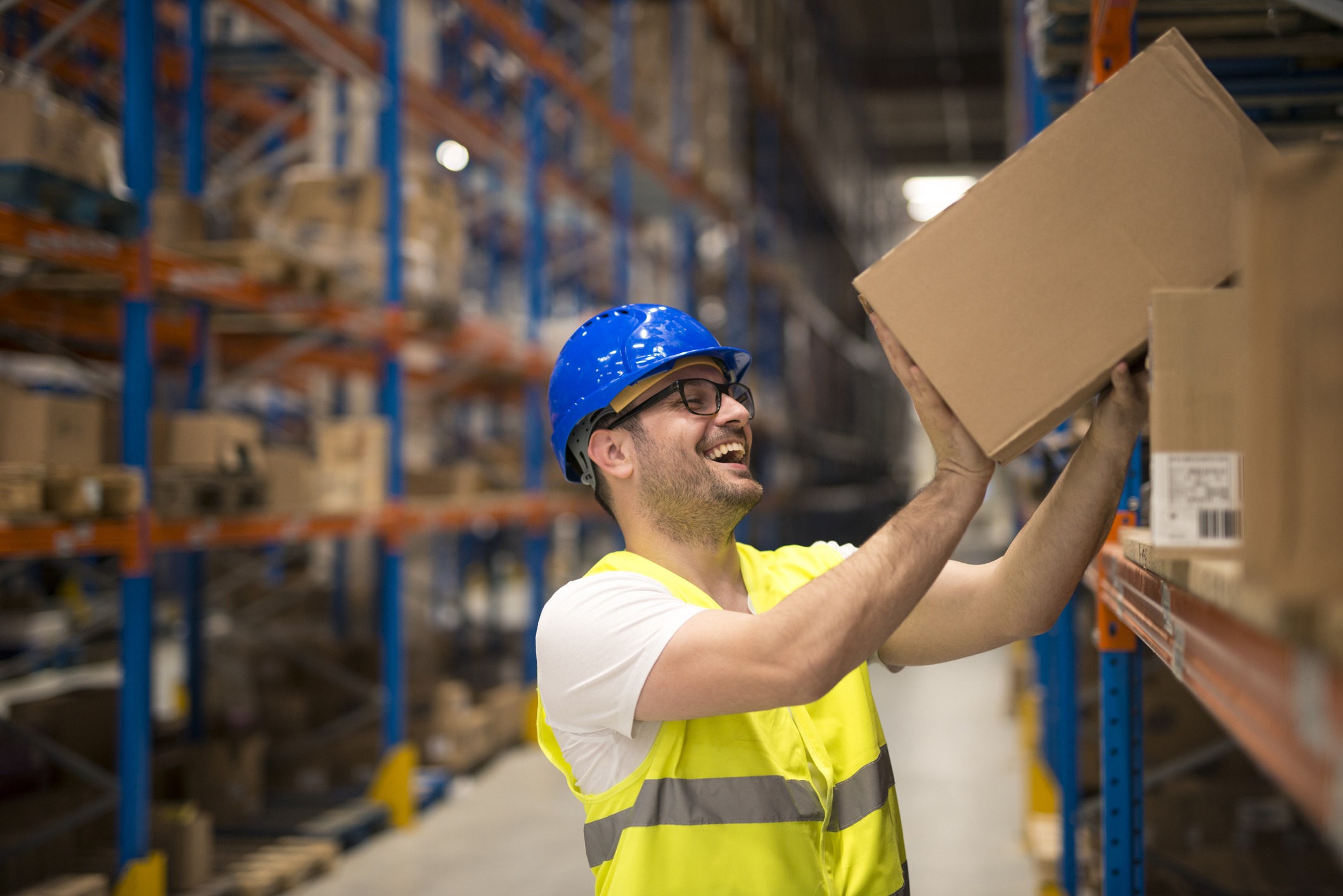 A smiling person wearing a blue helmet and safety vest lifts a box in a warehouse with tall shelves filled with goods in the background.