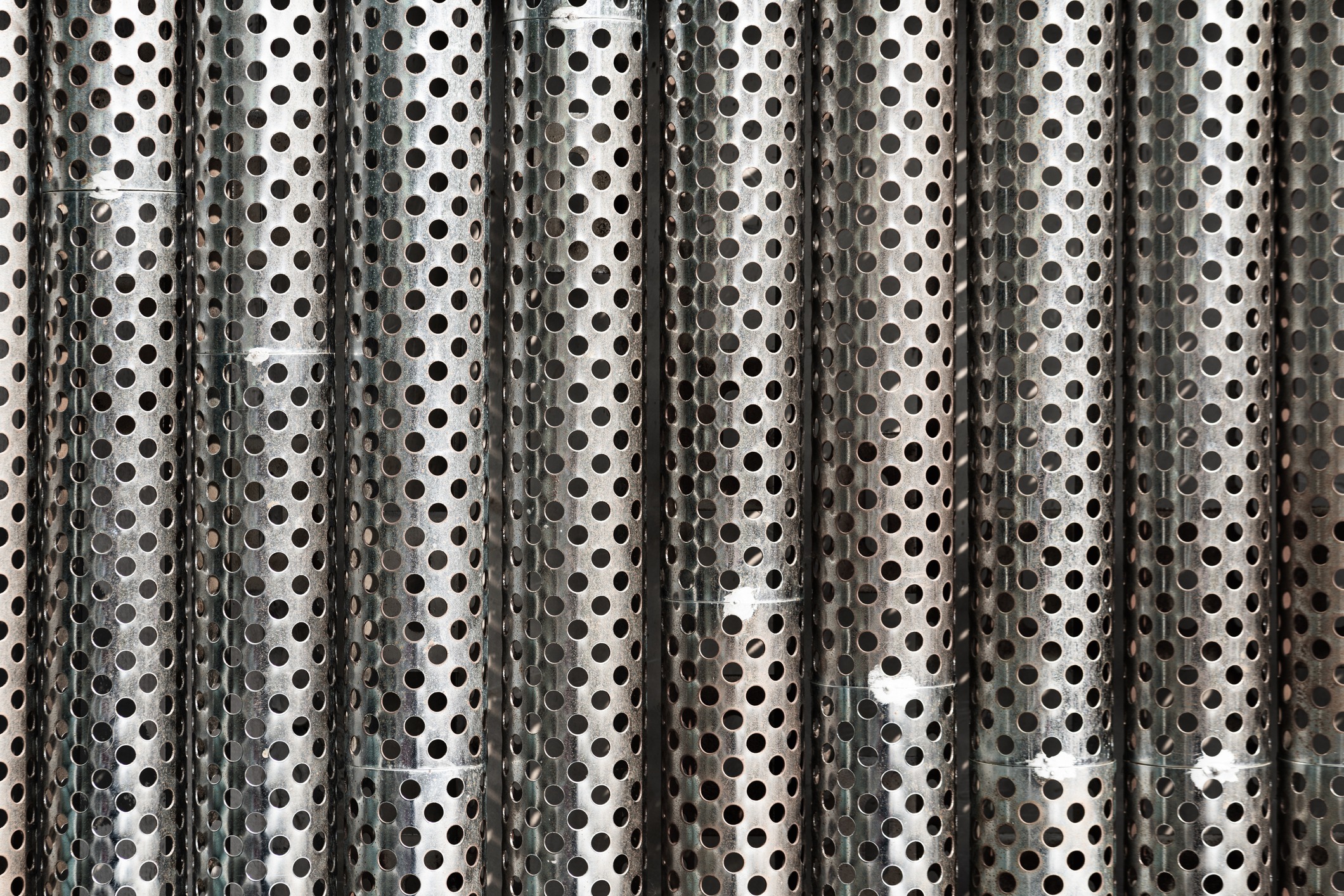 The image shows a close-up of vertical metal sheets with numerous circular holes, presenting an industrial or modern textured appearance with metallic tones.