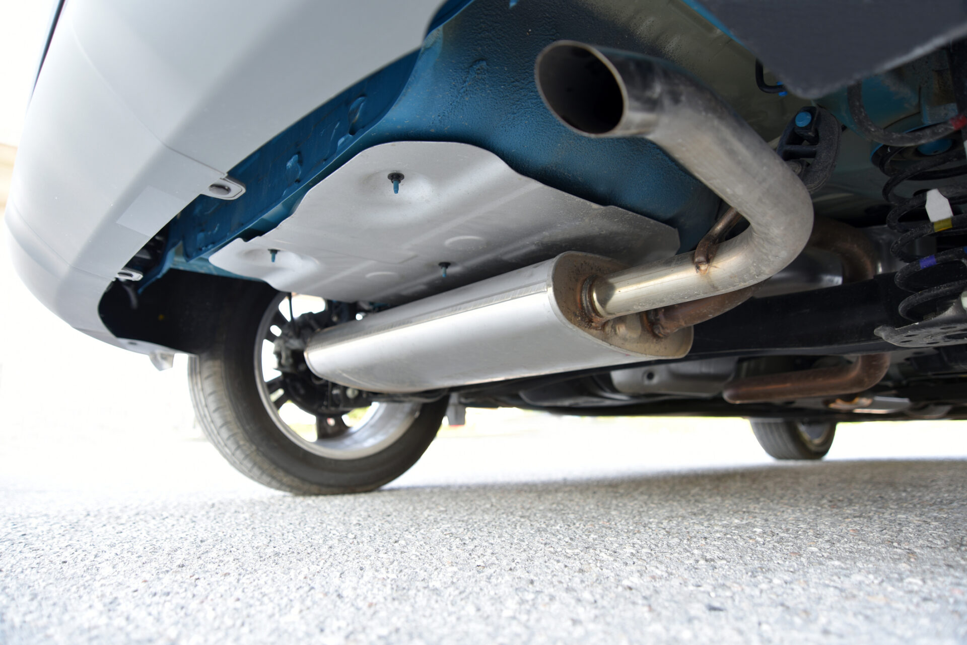 The image shows the underside of a vehicle, featuring a metal exhaust pipe and parts of the suspension system against an asphalt surface.