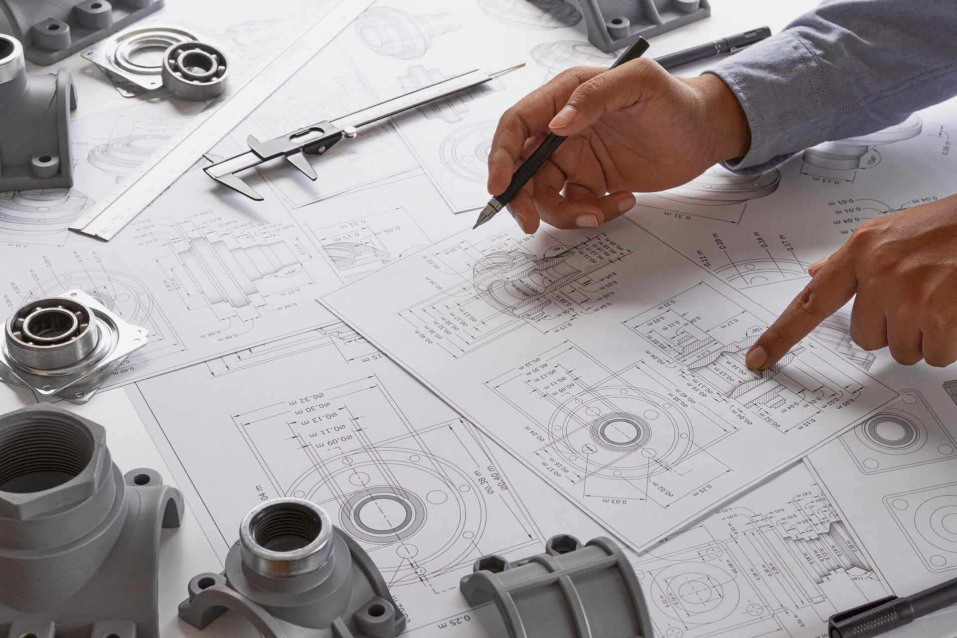 This photo shows a person with a pencil over mechanical engineering drawings surrounded by machine parts, vernier calipers, and rulers, indicating design or review work.