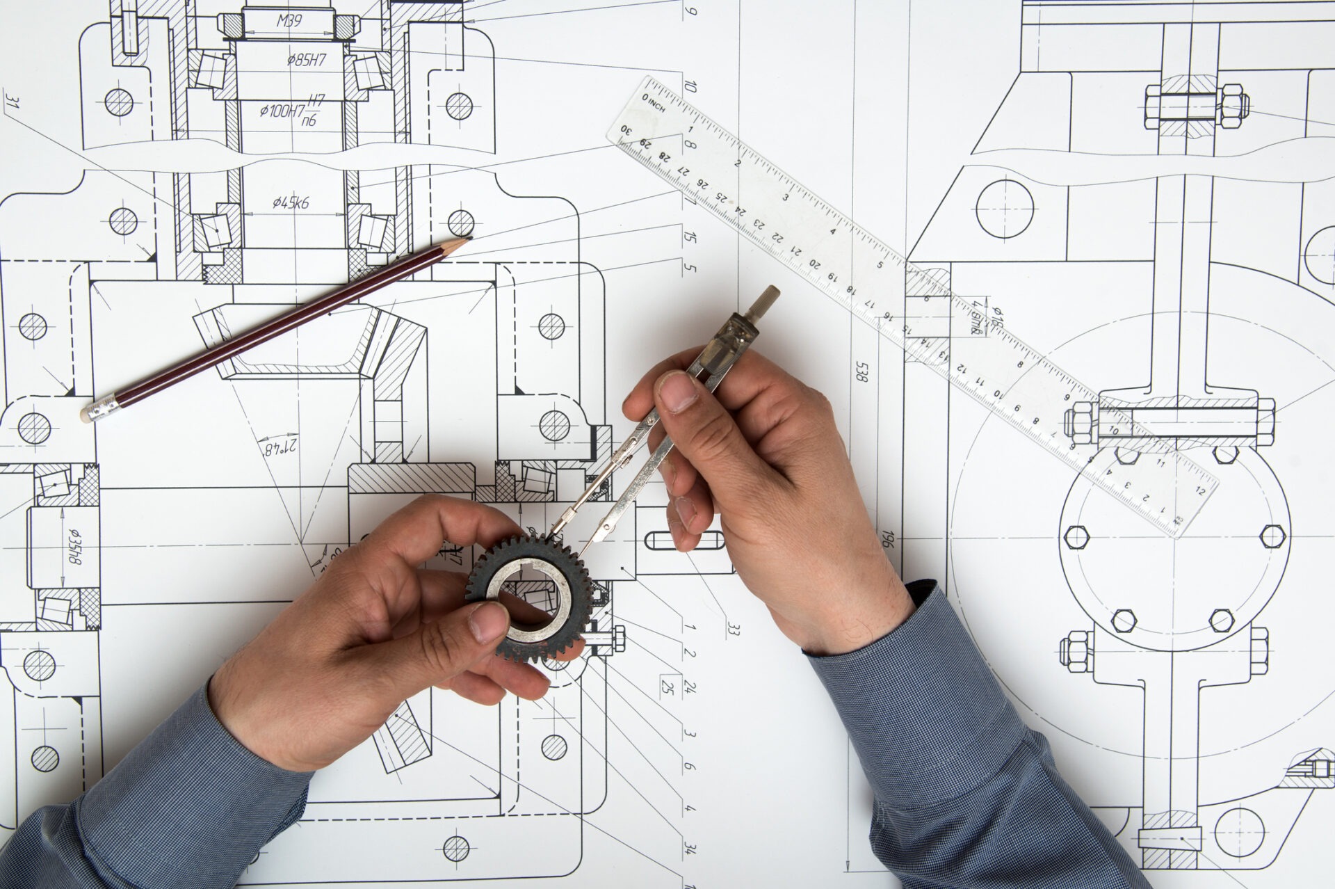 A person's hands are holding a vernier caliper and a gear over a detailed mechanical blueprint with a pencil and rulers nearby.