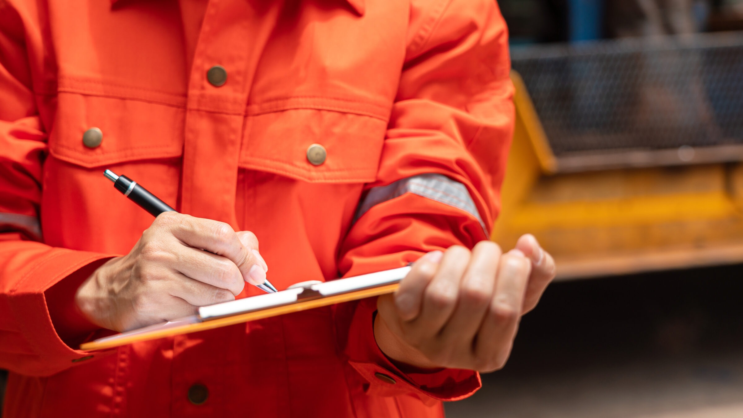 A person in a bright orange safety suit is writing on a clipboard with a pen, likely taking notes or conducting an inspection.