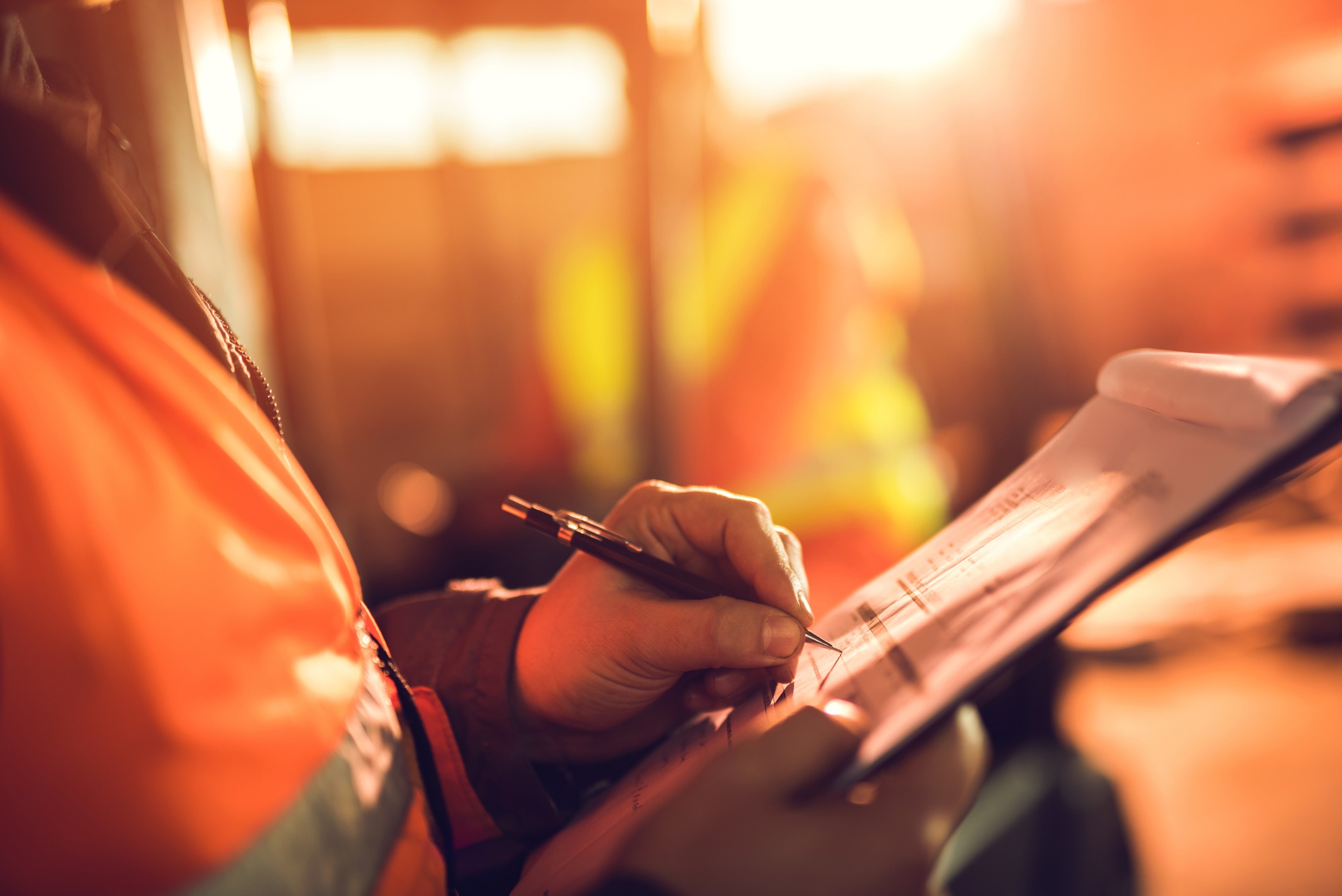 A person in a reflective orange vest writes on a clipboard, bathed in warm sunlight, suggesting an industrial or construction setting.