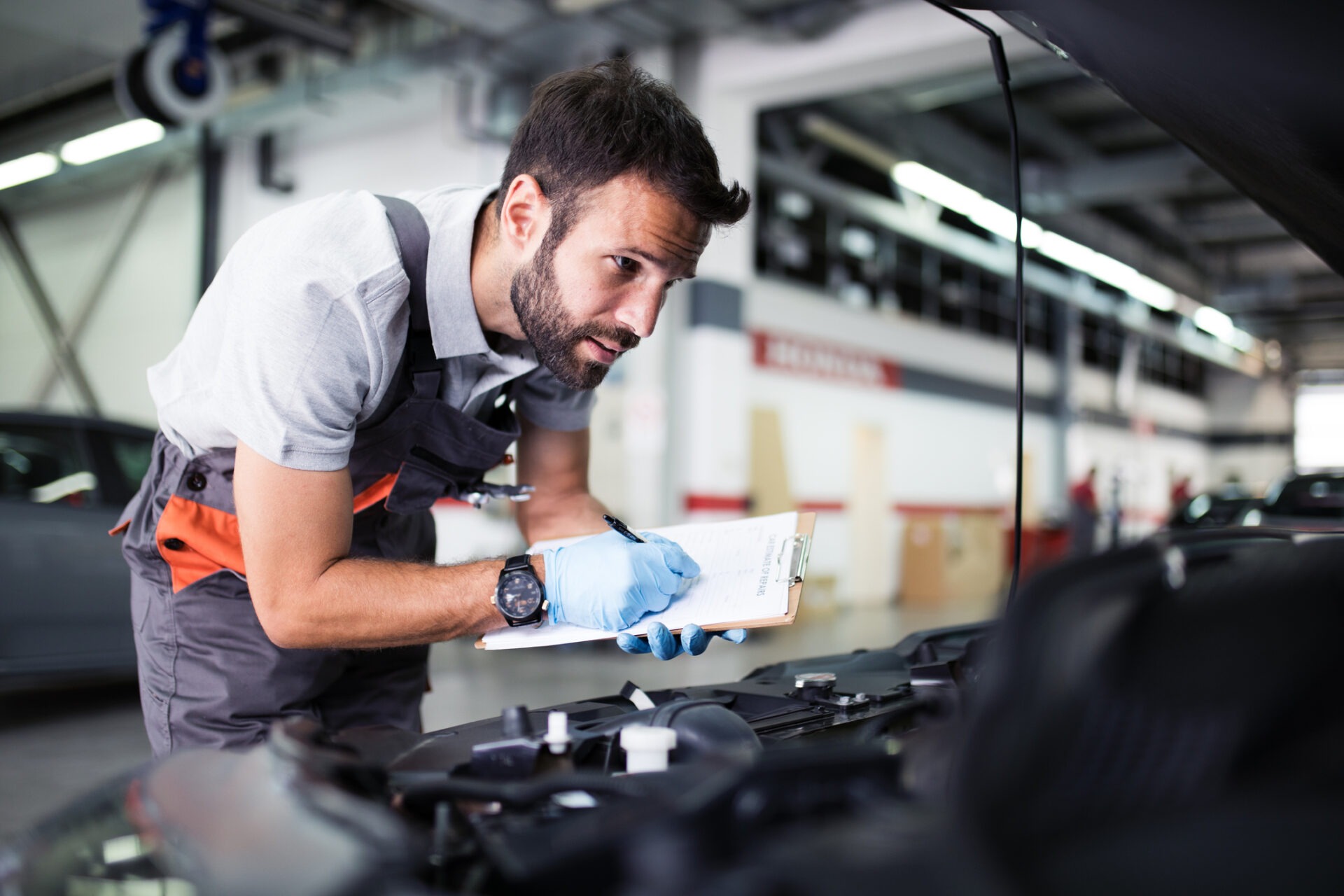 A person is inspecting a car engine while holding a checklist at an automotive workshop. They're focused, wearing gloves, and a wristwatch.